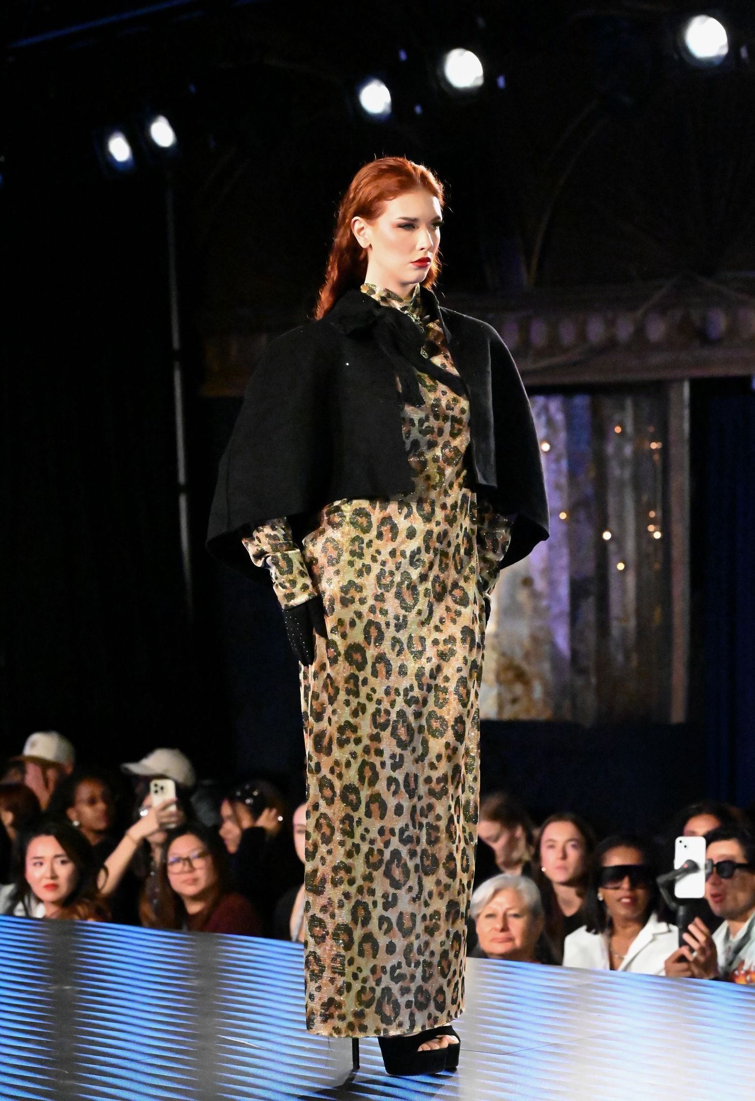 Model walks runway in leopard print dress and black turtleneck during fashion show with audience visible.