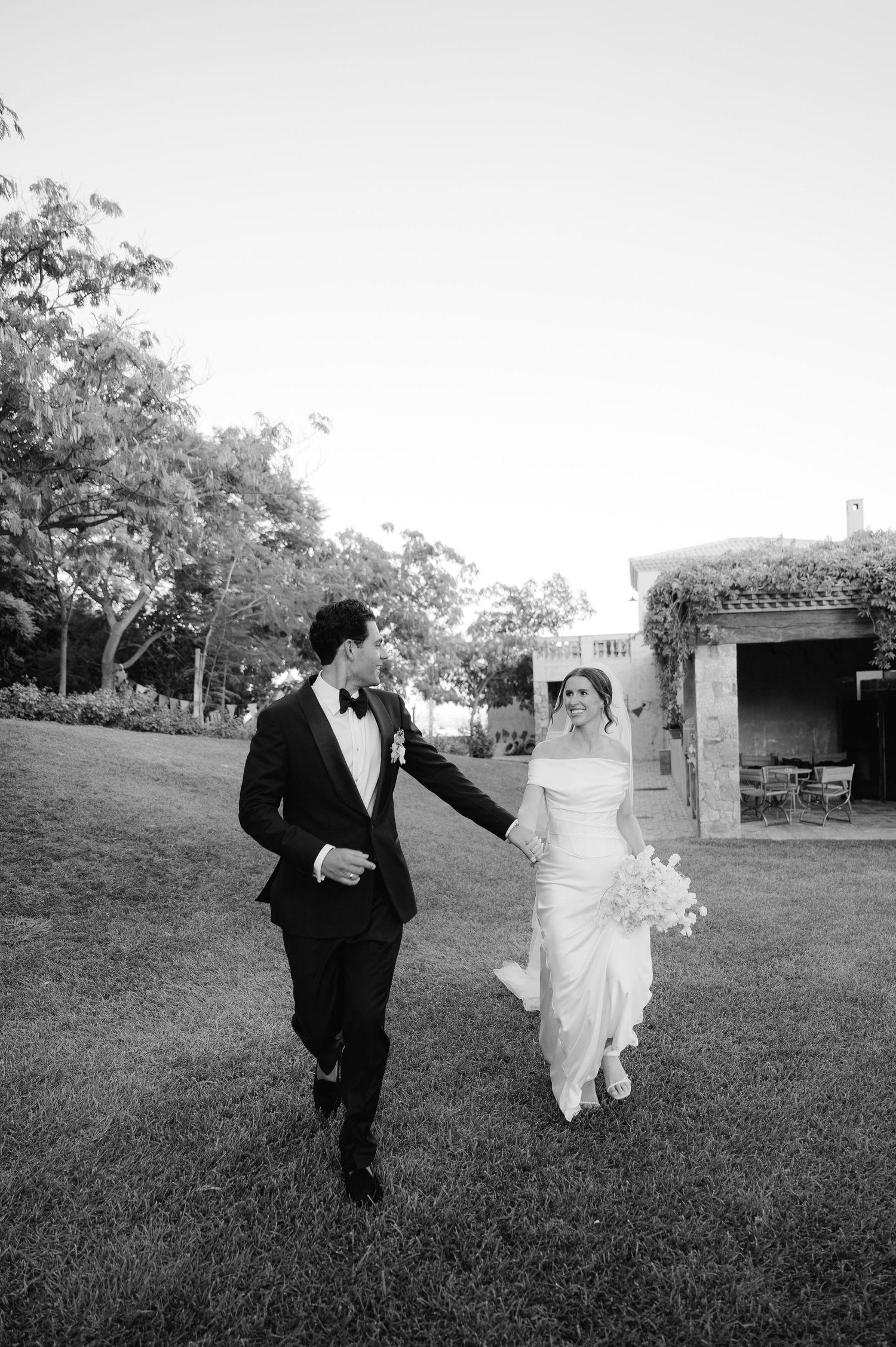 Newlyweds playfully run and dance together on a lush green lawn at their outdoor wedding venue.