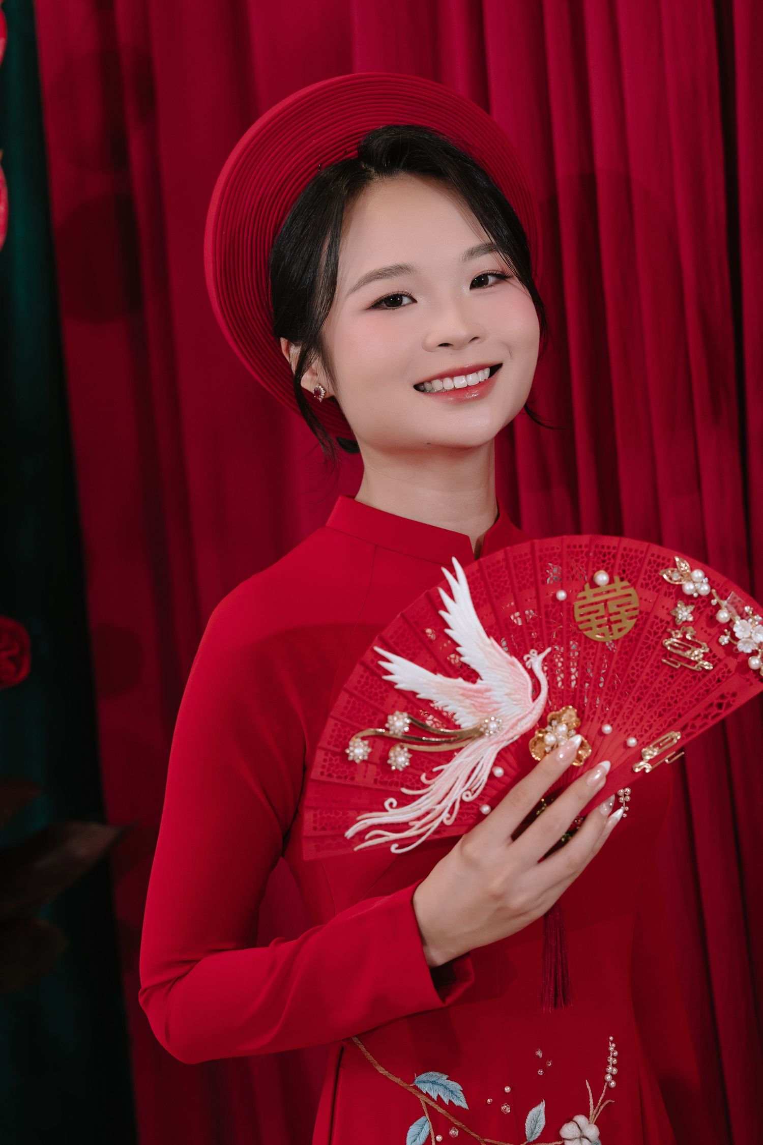 Smiling Vietnamese bride in red ao dai holds embroidered fan with white phoenix design.
