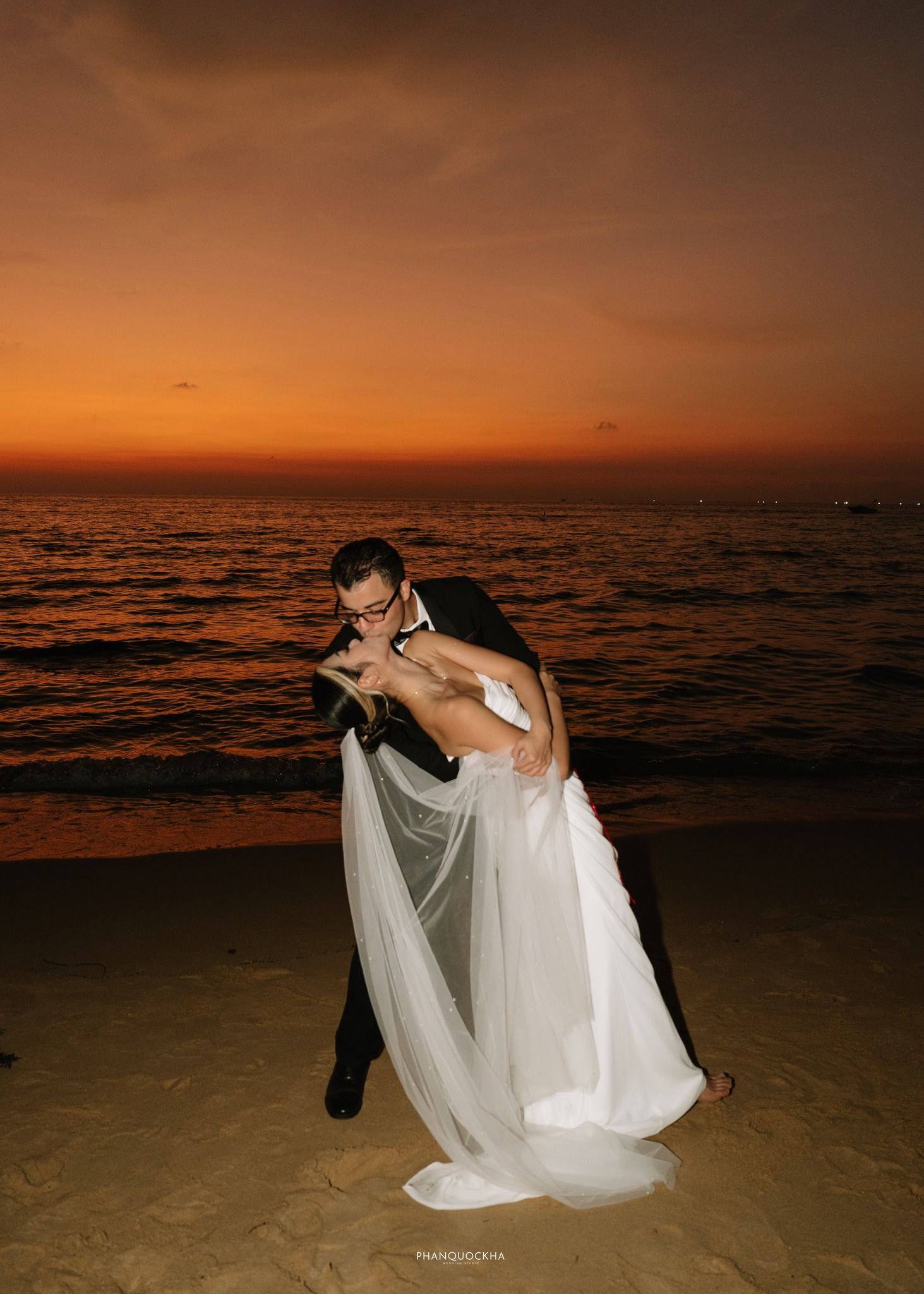 Romantic bride and groom sharing a dip kiss on sandy beach at stunning orange sunset.