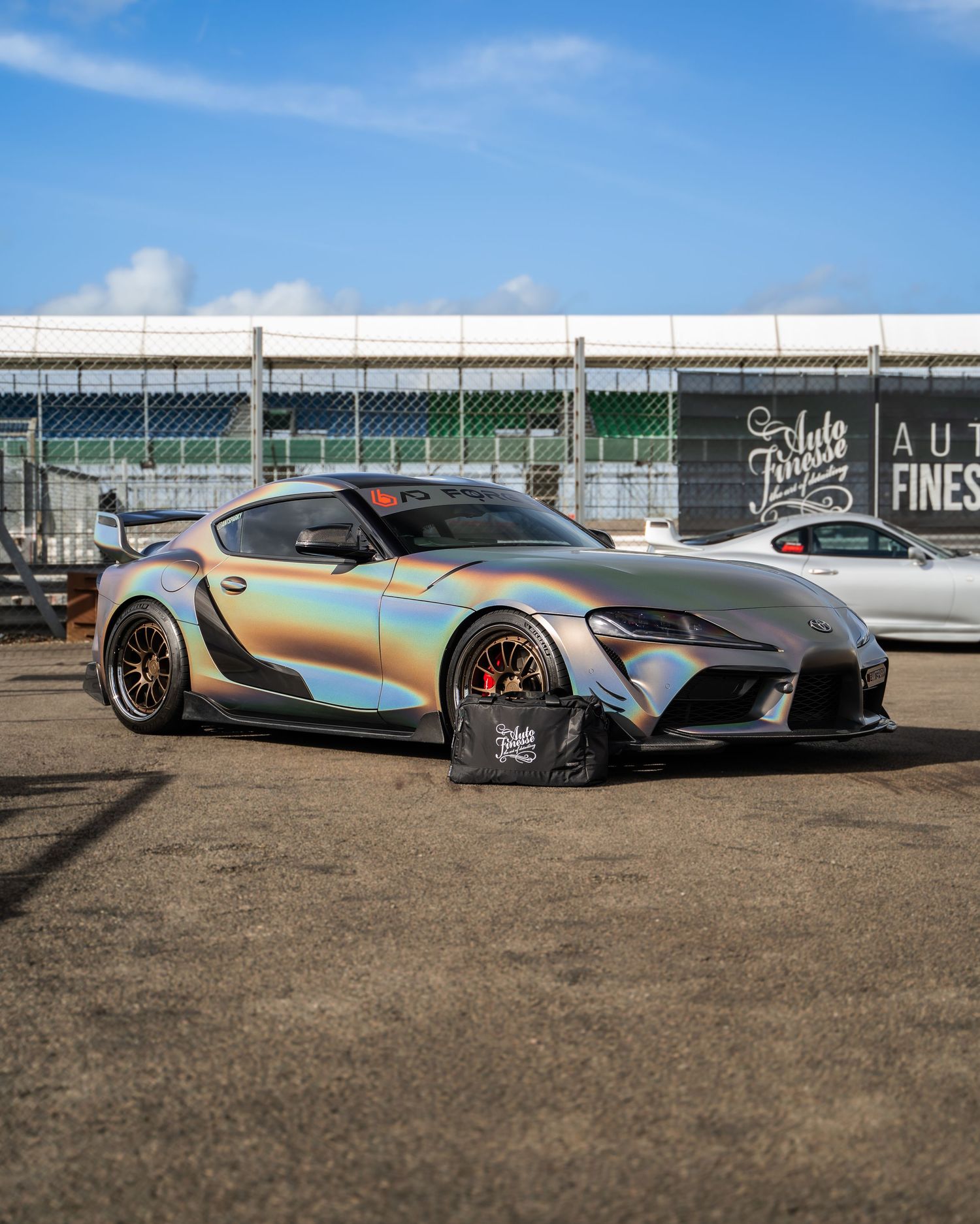 Iridescent Toyota Supra sports car with custom body kit parked in front of industrial building.