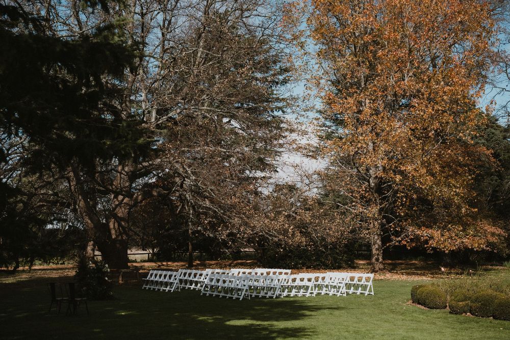 White chairs set up in rows on a lawn for an outdoor event with fall trees in the background.
