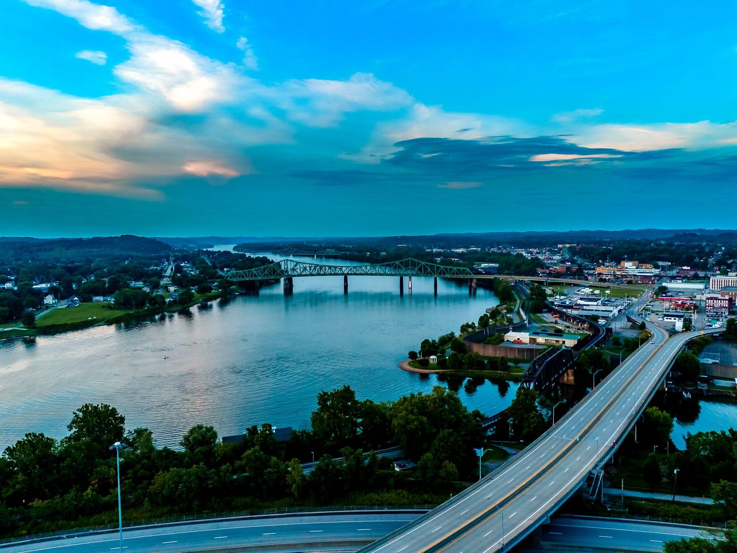 Panoramic aerial view of a highway bridge spanning across a serene blue river on a sunny day.
