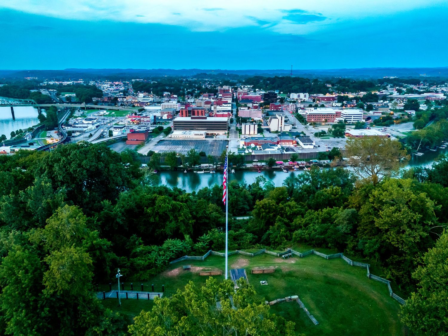 Aerial view of a small town cityscape with green trees in foreground and buildings nestled along a river under blue sky.