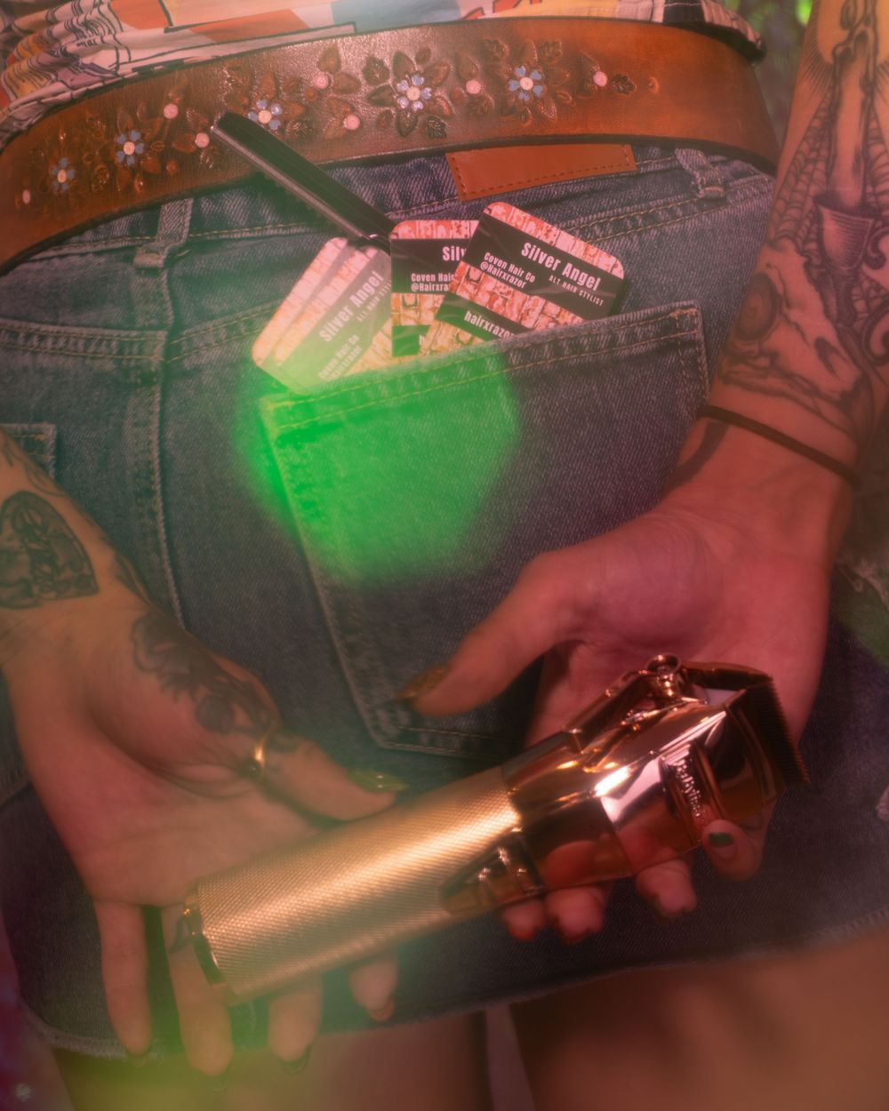 Close-up of tattooed hands holding gold hair clippers with business cards in denim jeans pocket.
