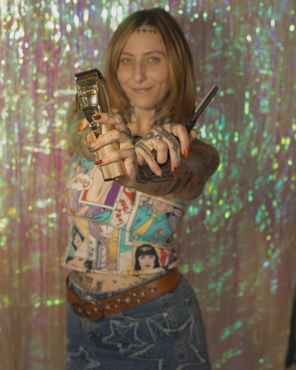 Tattooed woman pointing gold hair clippers at camera against an iridescent holographic backdrop.