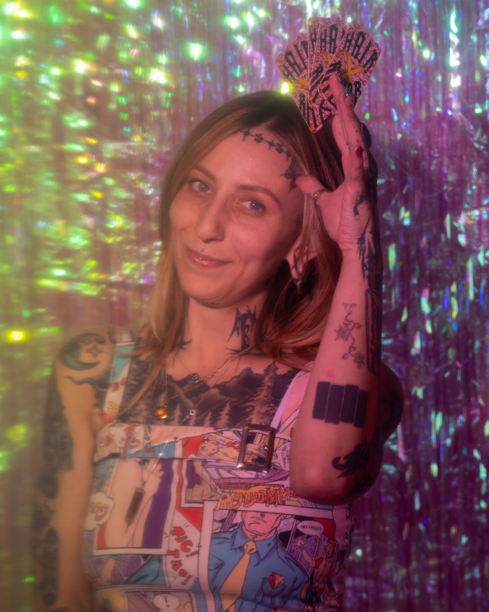 Tattooed woman posing against a sparkling green iridescent backdrop wearing a comic book print top.