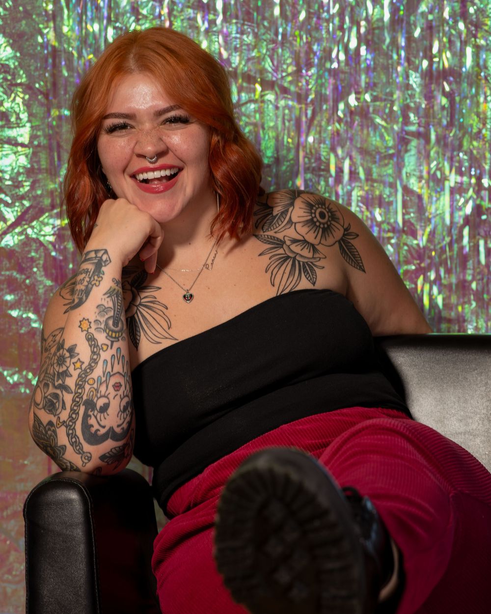 Redhead tattooed woman laughing in black top and red pants against a holographic green backdrop.