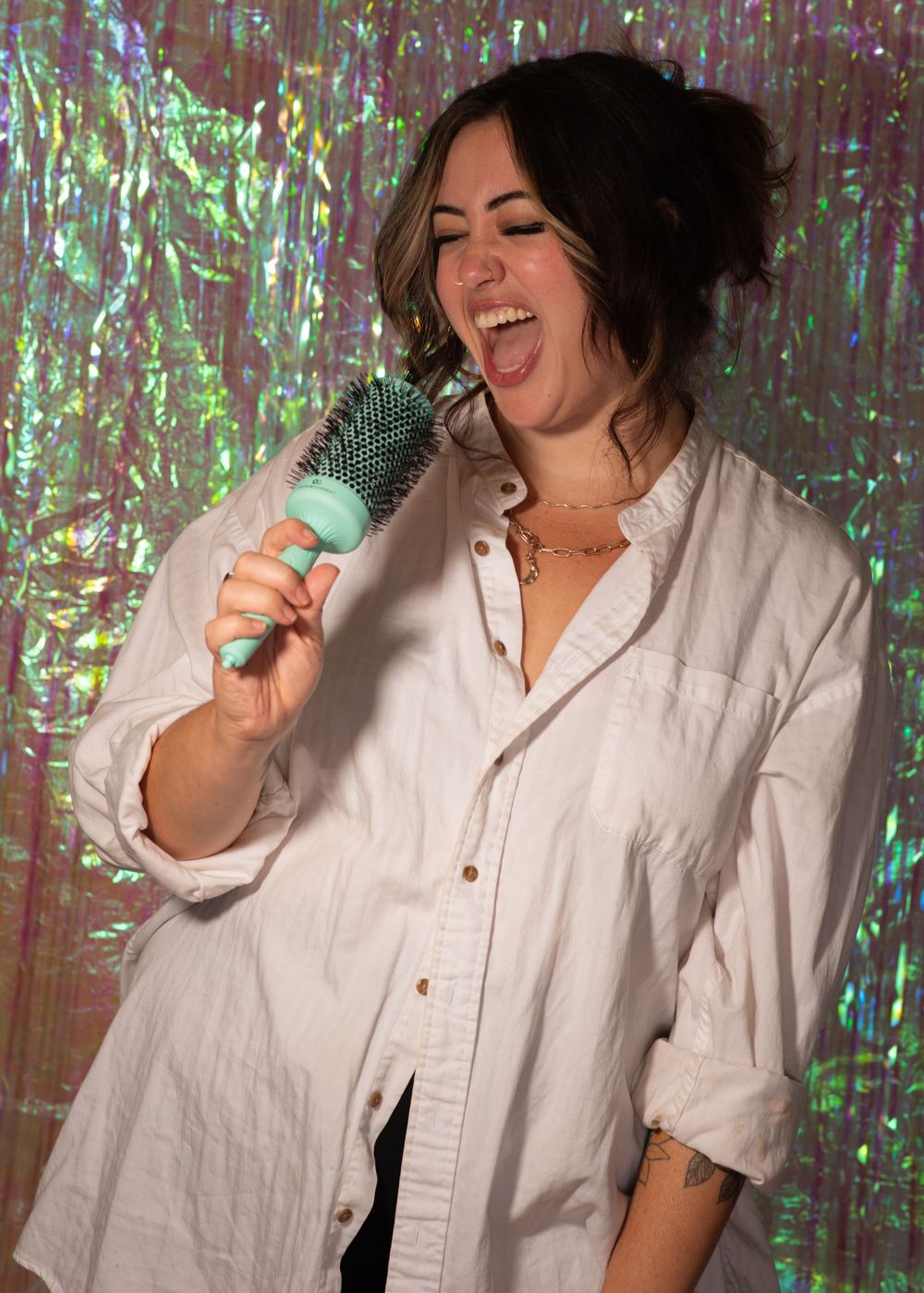Laughing woman in white shirt holding a teal round brush like a microphone against holographic backdrop.