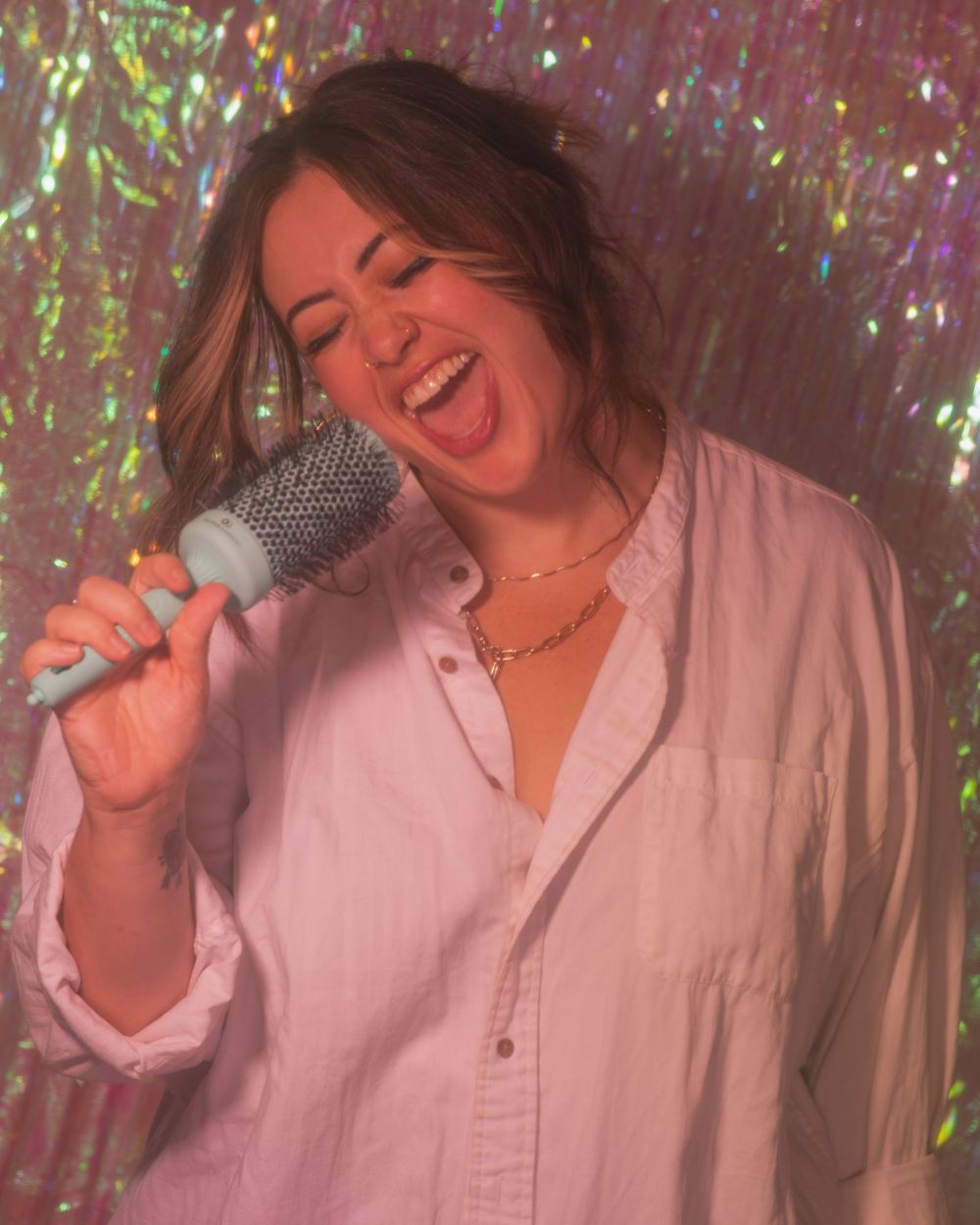 Joyful woman singing into a teal round brush against a sparkling holographic glitter curtain background.