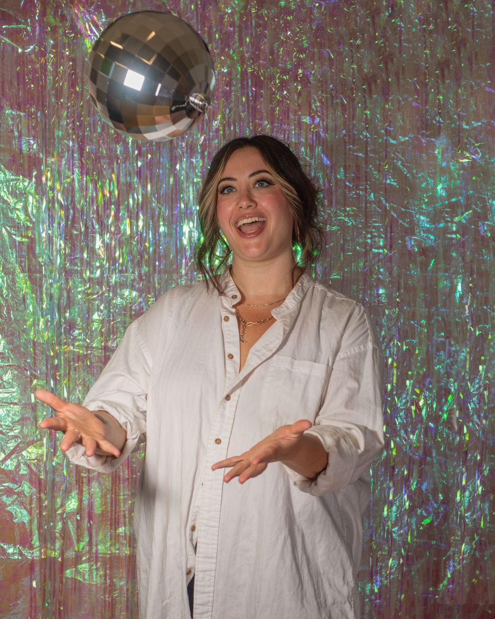 Woman in white shirt laughing and tossing a disco ball against a holographic iridescent backdrop.
