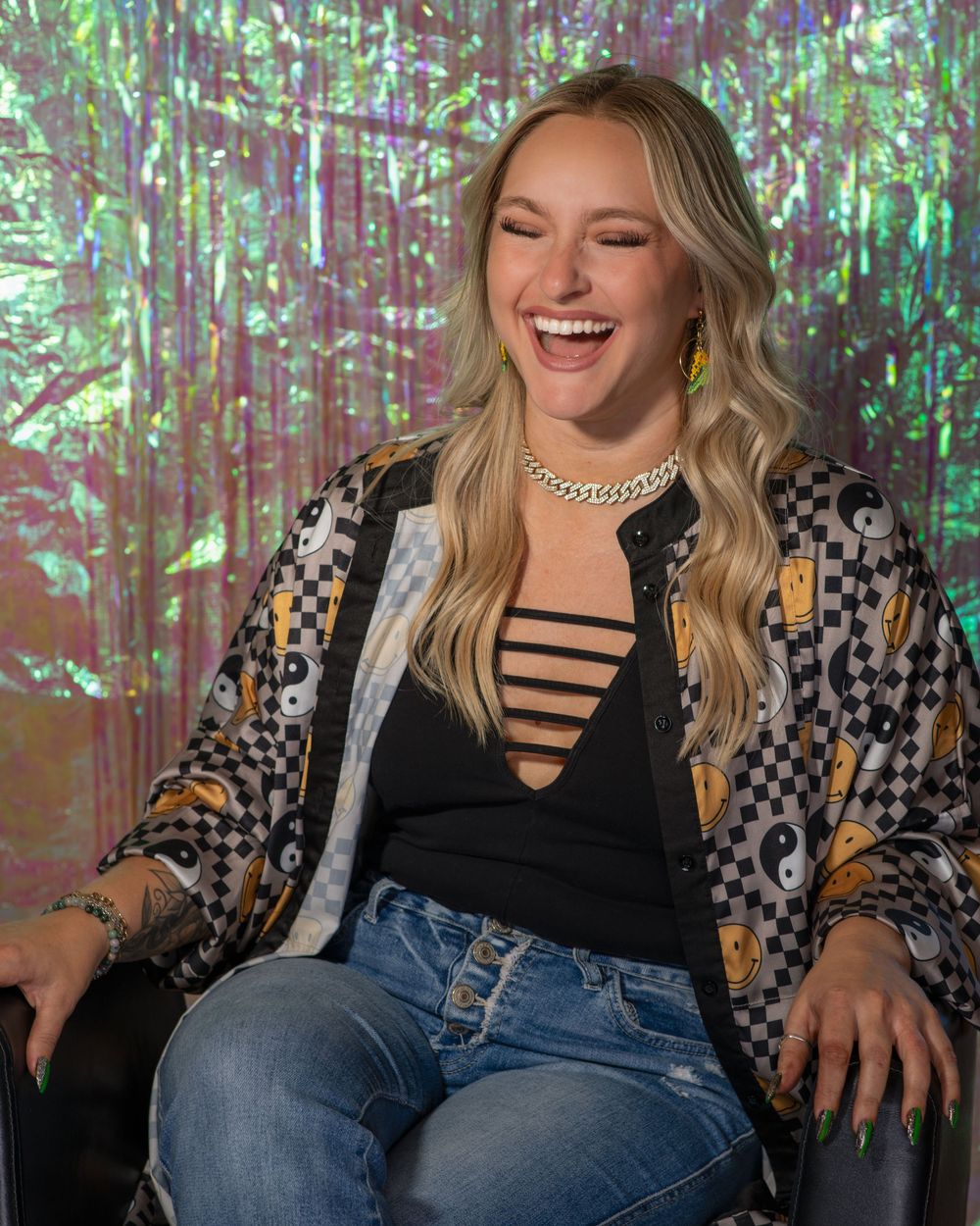 Blonde woman laughing in patterned jacket and jeans seated against a colorful holographic background.