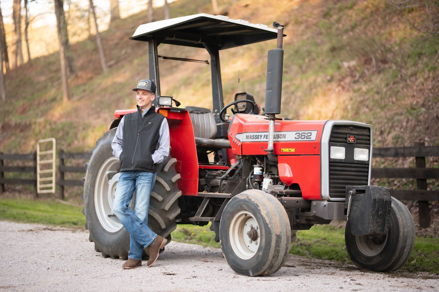 A red Massey Ferguson tractor is parked on a gravel path with a person leaning against it in a rural farm setting.