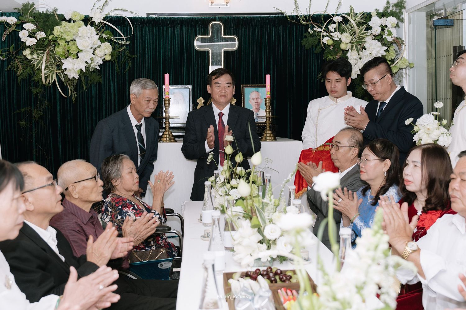 Guests applaud at Vietnamese Christian wedding reception with cross backdrop, floral arrangements, and traditional ceremony table setting.