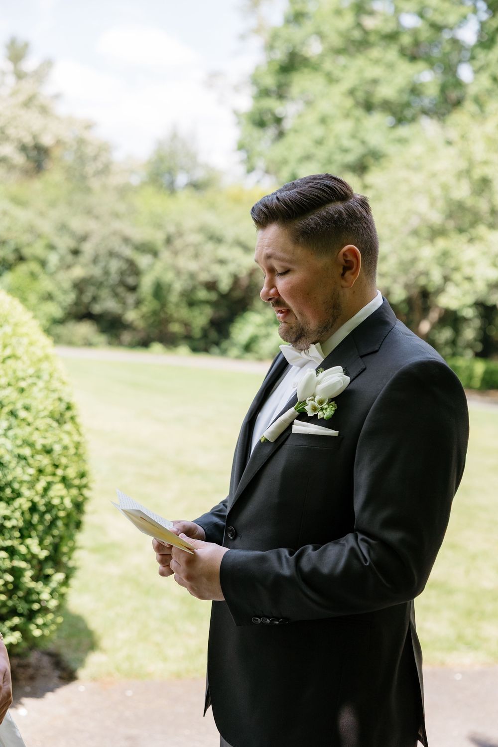 Groomsman in black tuxedo reads emotional letter outdoors in garden setting.
