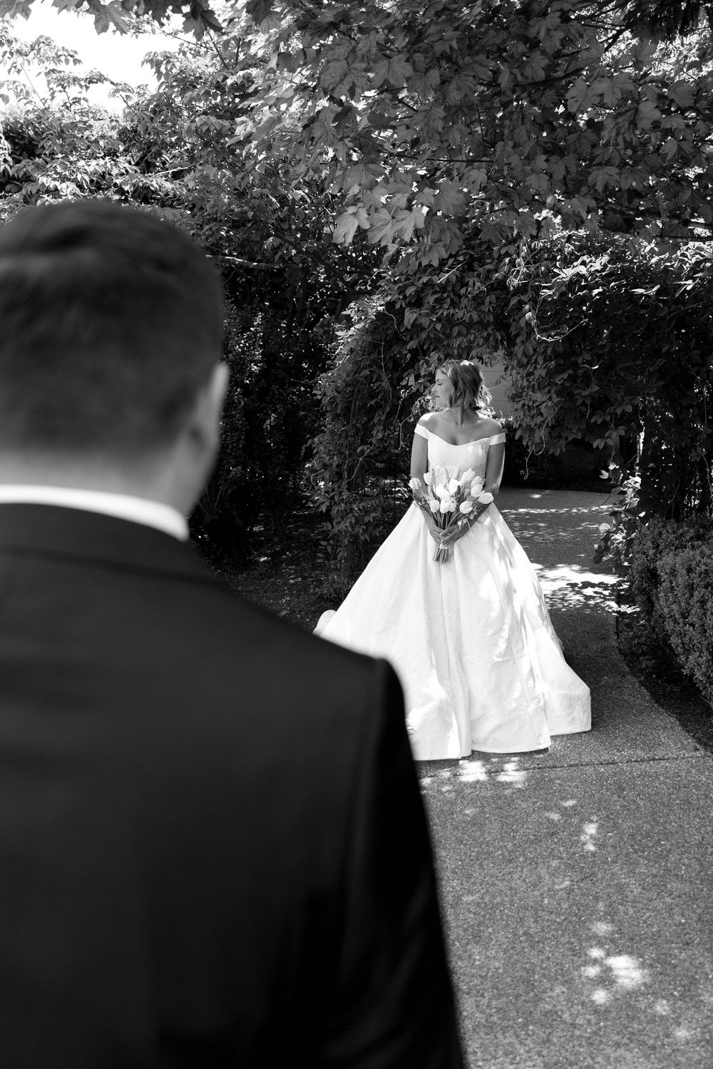 A bride in a flowing white wedding dress twirls gracefully in a garden setting as a suited figure watches from the side.
