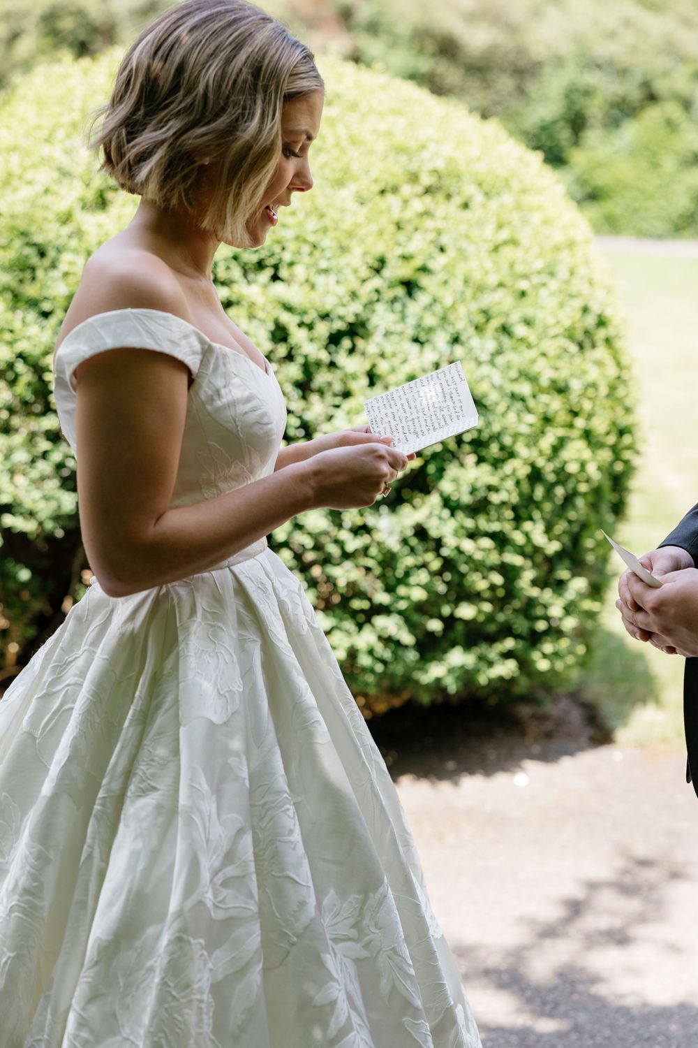 Someone in a white wedding dress reads from a paper in an outdoor garden setting.