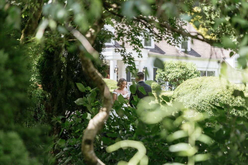 A white house peeks through lush green foliage and tree branches in a garden setting.