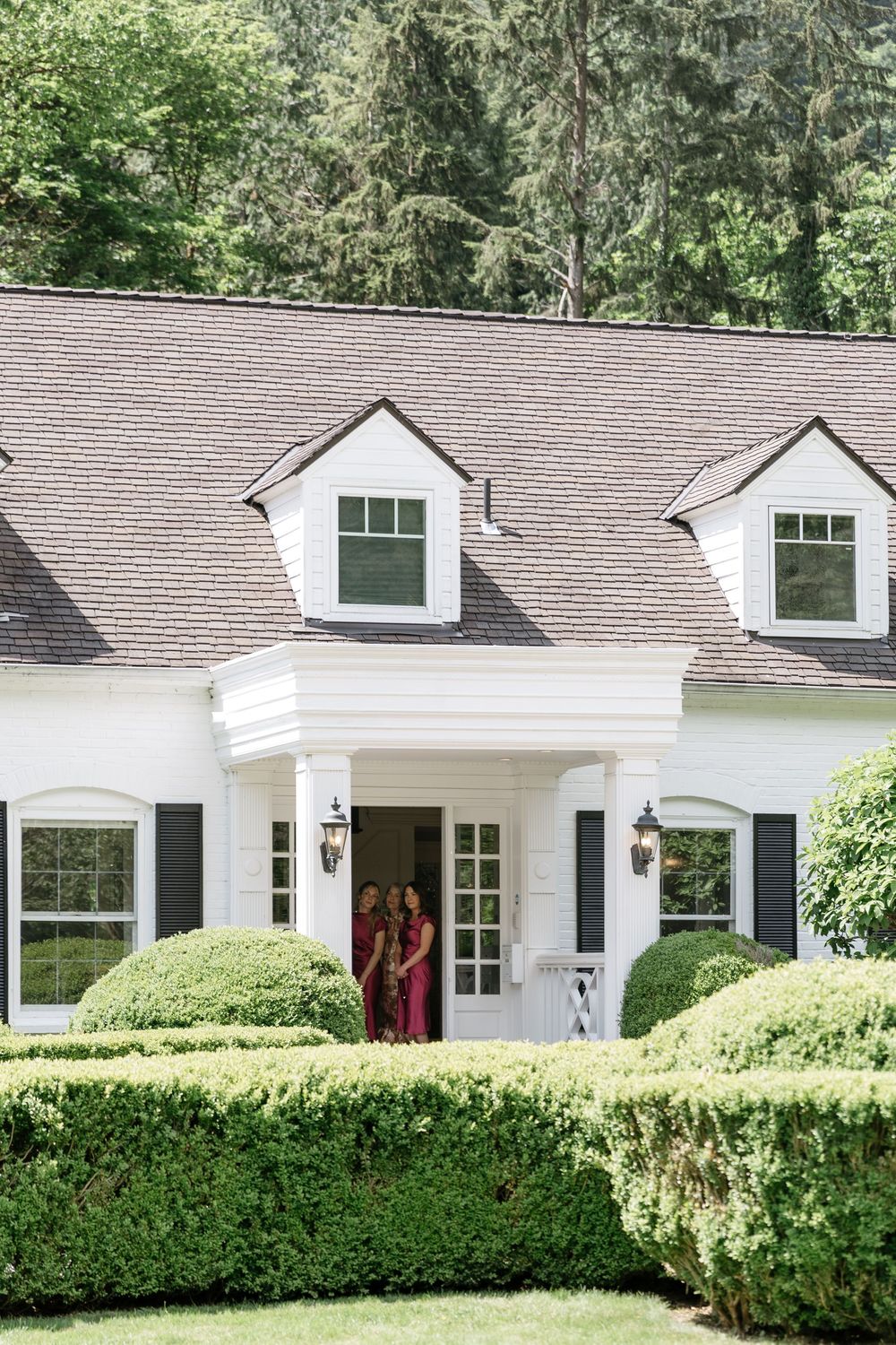 A white ranch-style house with black shutters and manicured hedges sits against a wooded backdrop.
