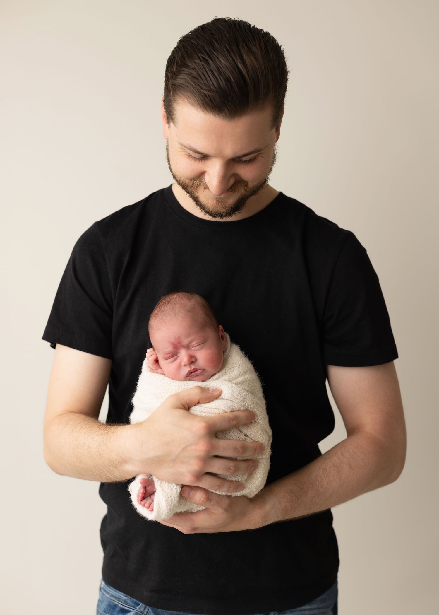 A person wearing a black t-shirt holds a swaddled newborn baby in a series of tender poses against a light backdrop.