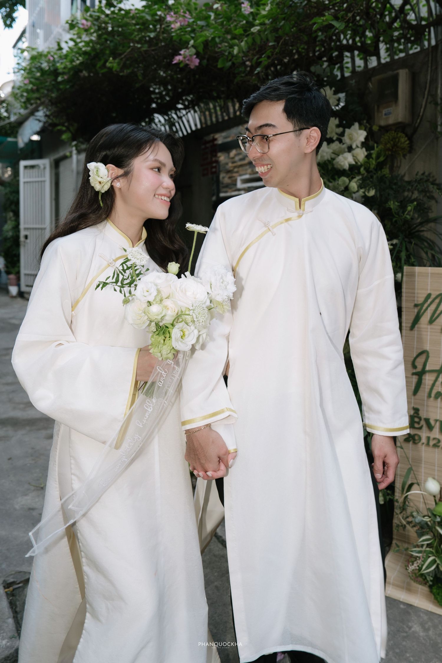 Vietnamese bride and groom in white ao dai with gold trim hold hands and exchange smiles.