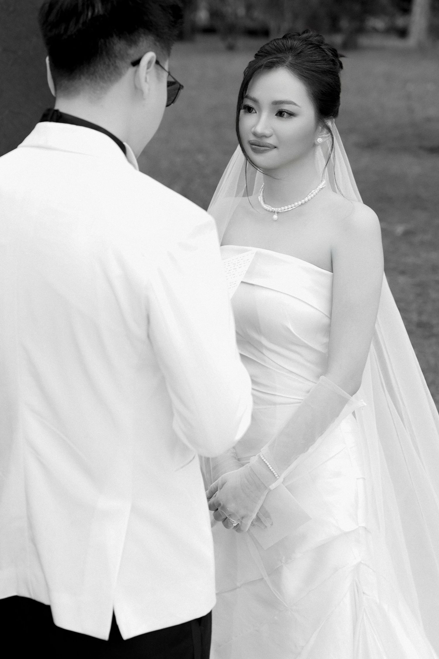 Black and white portrait of Asian bride in strapless gown and veil holding grooms hands outdoors.