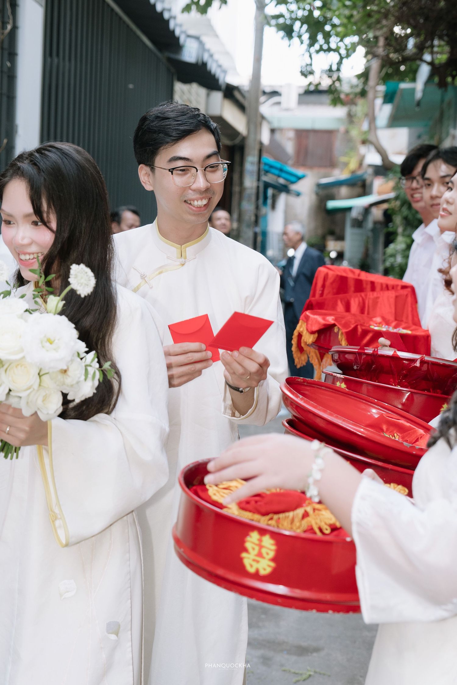 Young man in ao dai holds red envelopes beside bride during Vietnamese wedding ceremony.