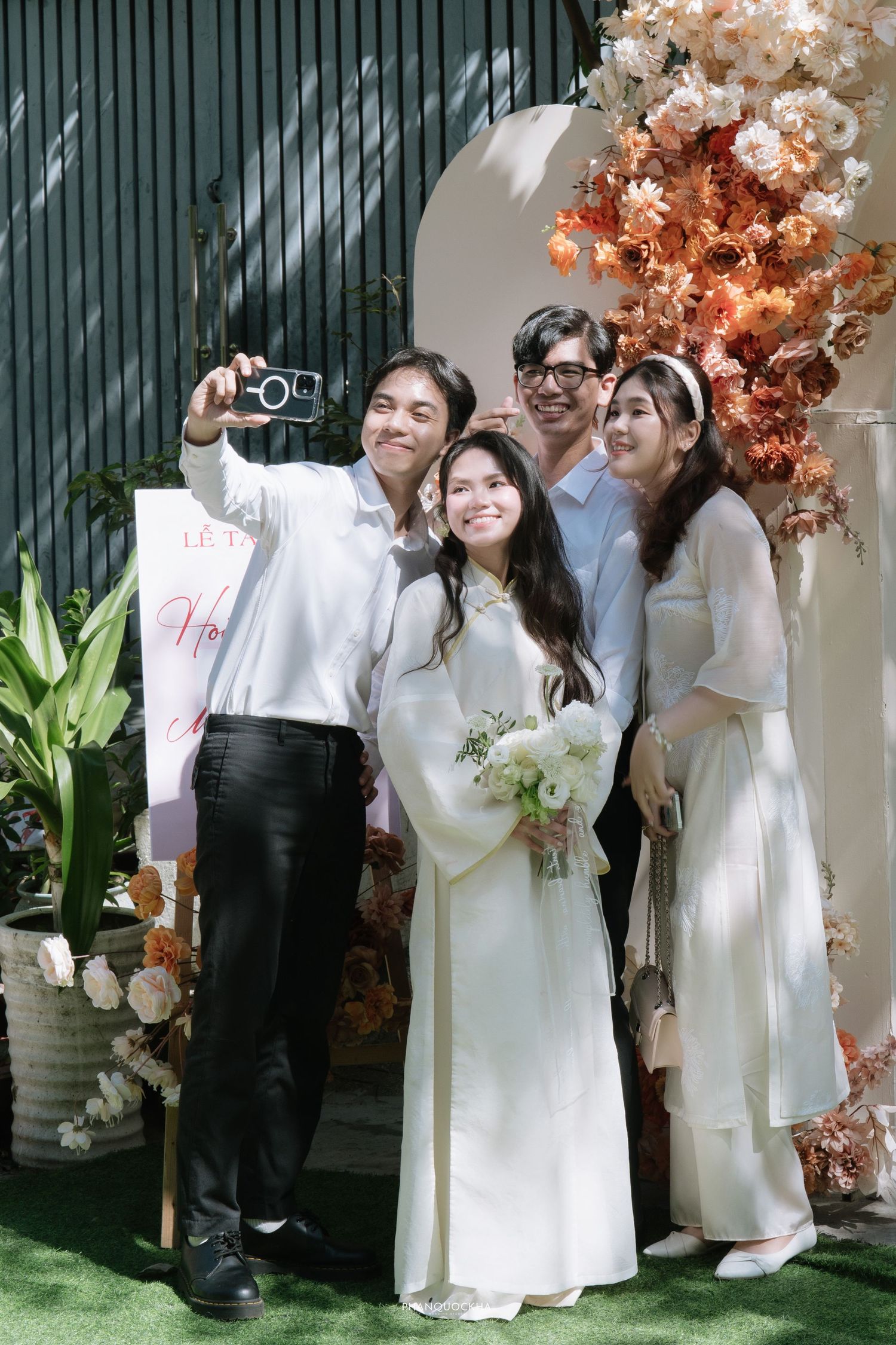 Wedding guests taking selfie with bride in white ao dai at outdoor Vietnamese wedding ceremony.