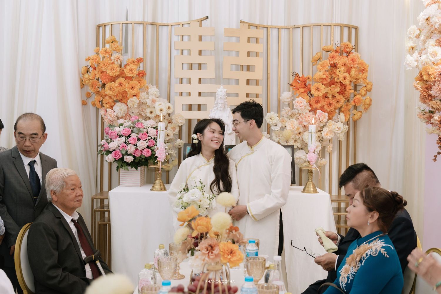Vietnamese wedding couple in white ao dai smile together at decorated reception table with guests.