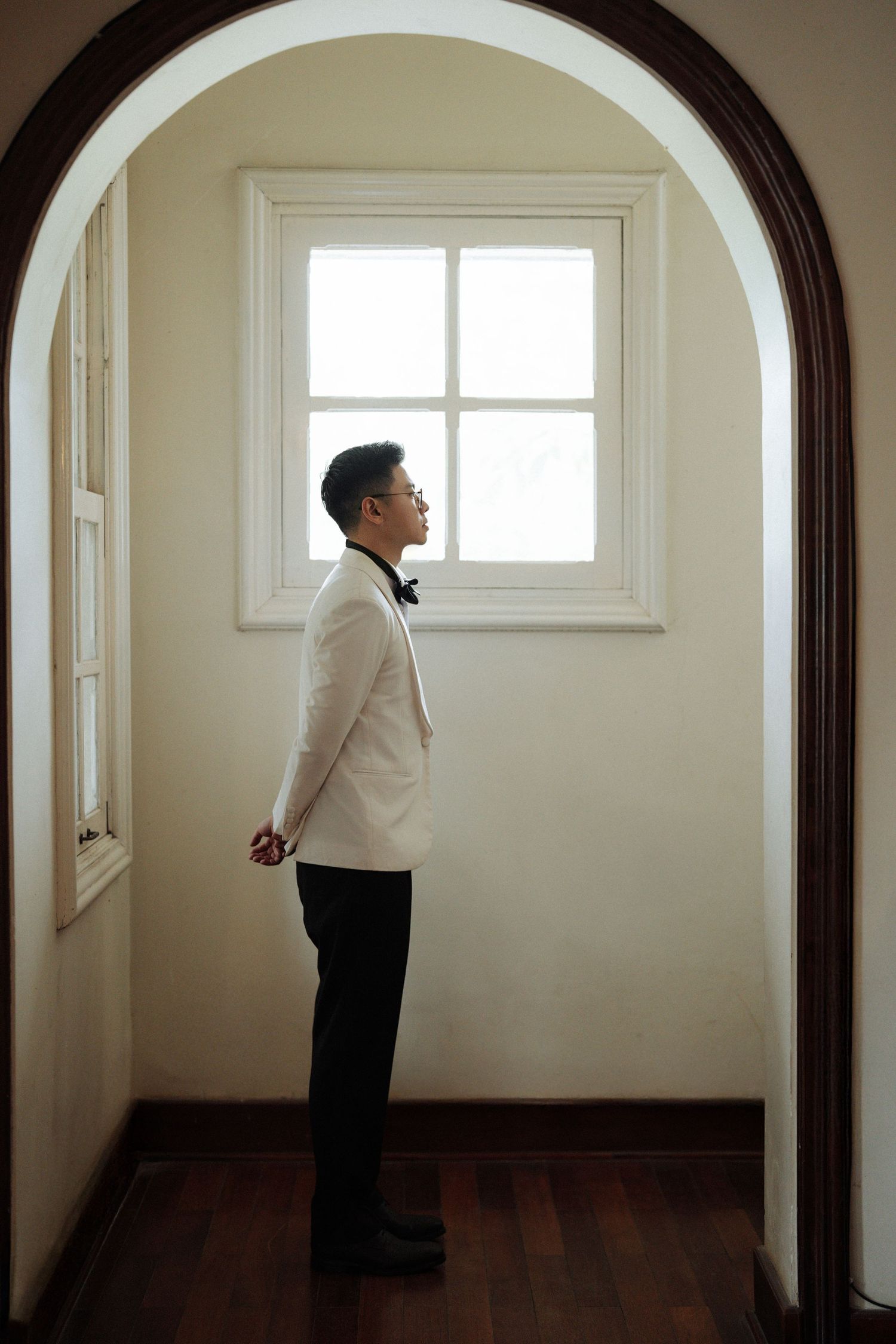 Dapper groom in white tuxedo jacket stands pensively beneath an arched doorway by a window.
