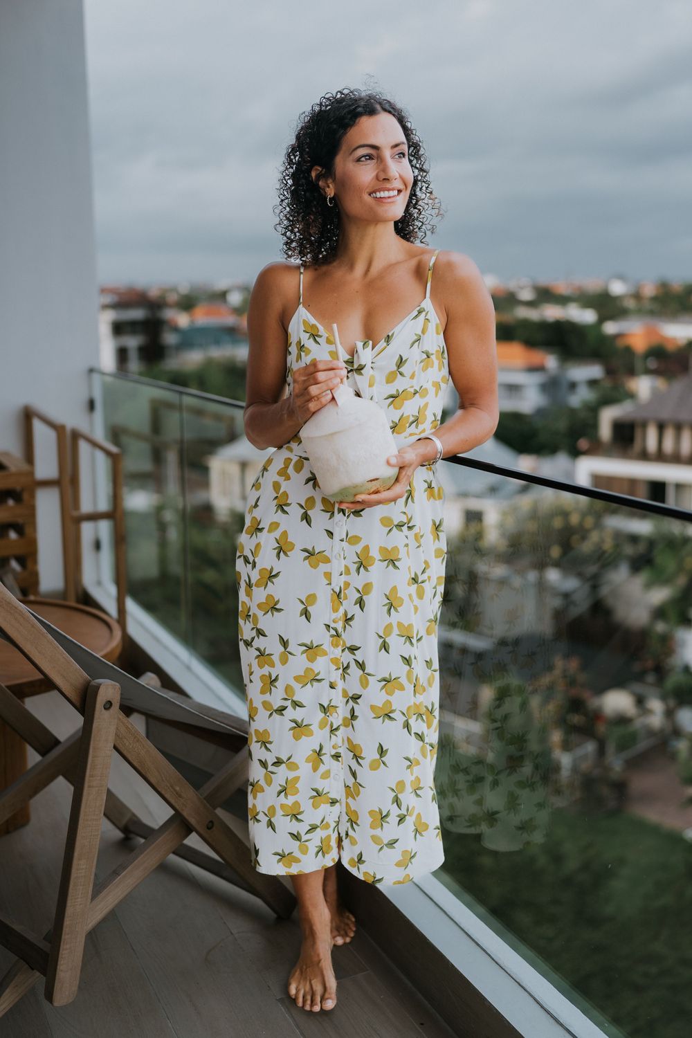 Someone in a white floral dress poses on an outdoor balcony with city views.