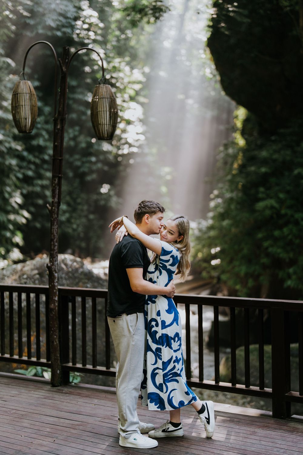 A couple embraces in a romantic pose on a wooden deck with forest backdrop and hanging lanterns.