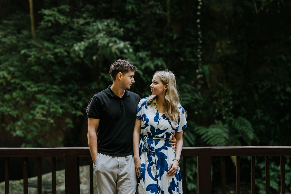 A couple stands together on a wooden boardwalk with lanterns and forest scenery.