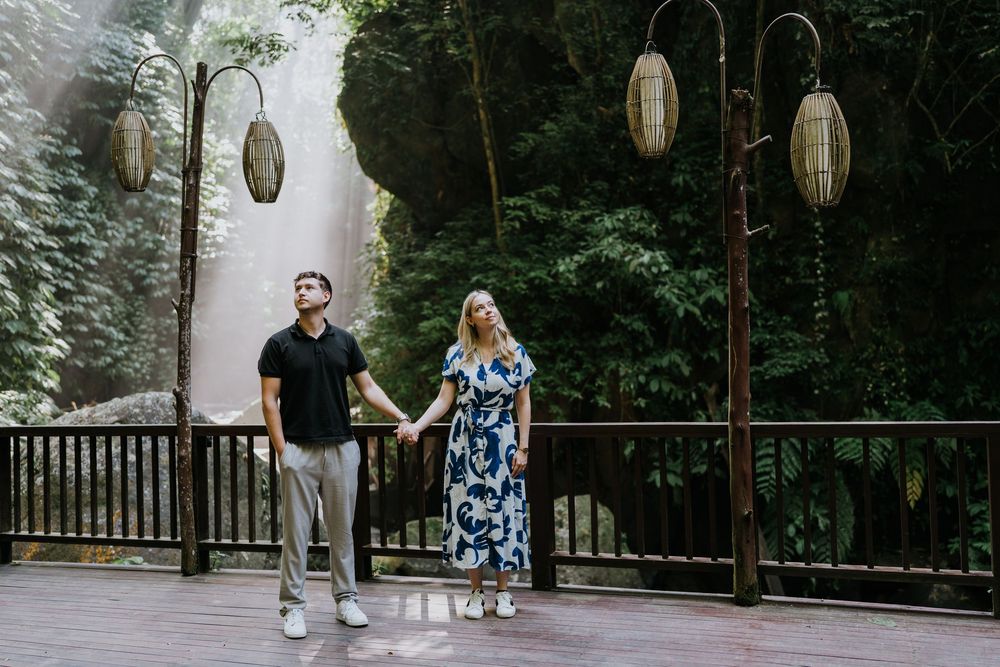 A couple walks together on a wooden deck surrounded by forest and hanging lanterns.