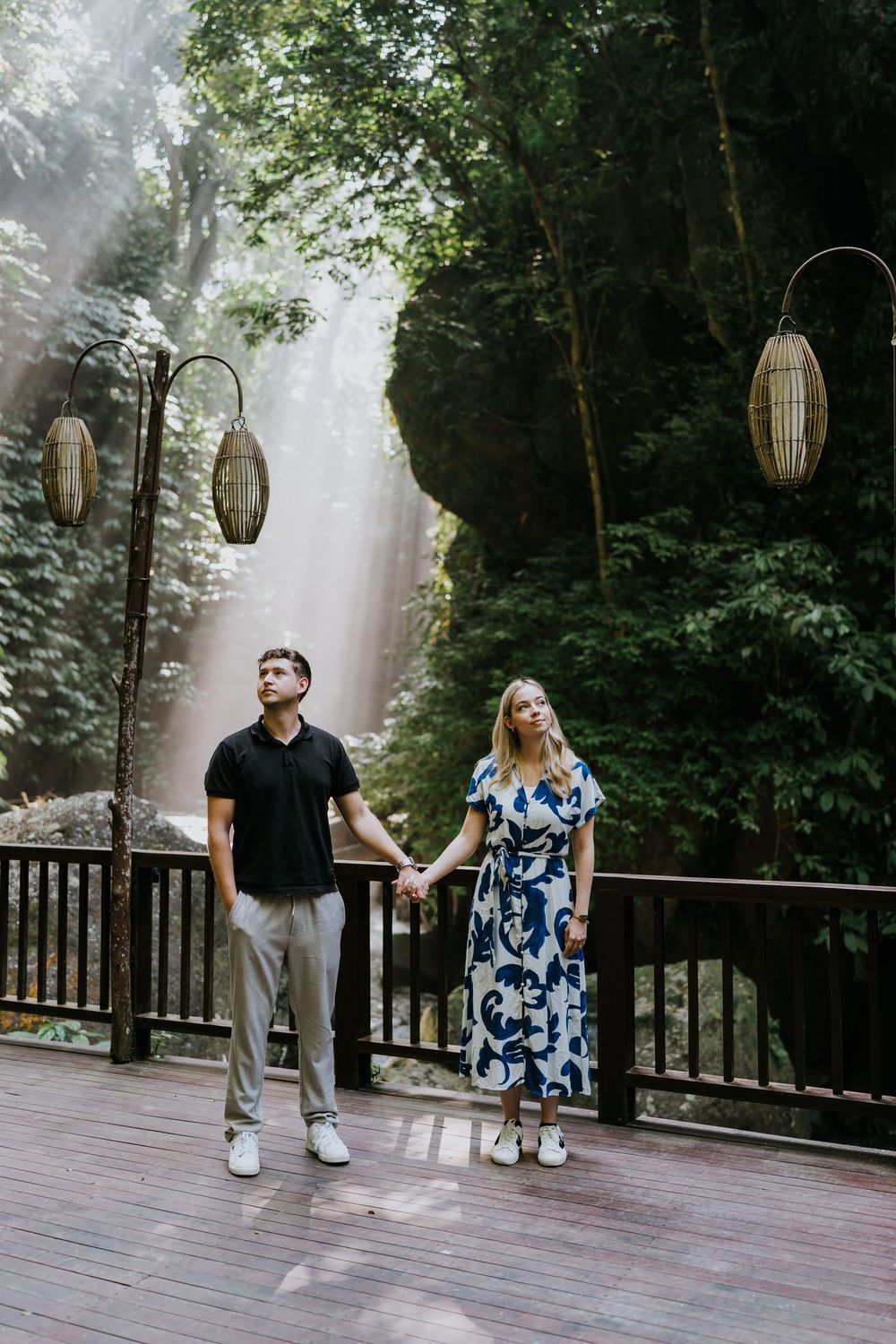 A couple holds hands on a wooden deck with misty forest and hanging lanterns in the background.