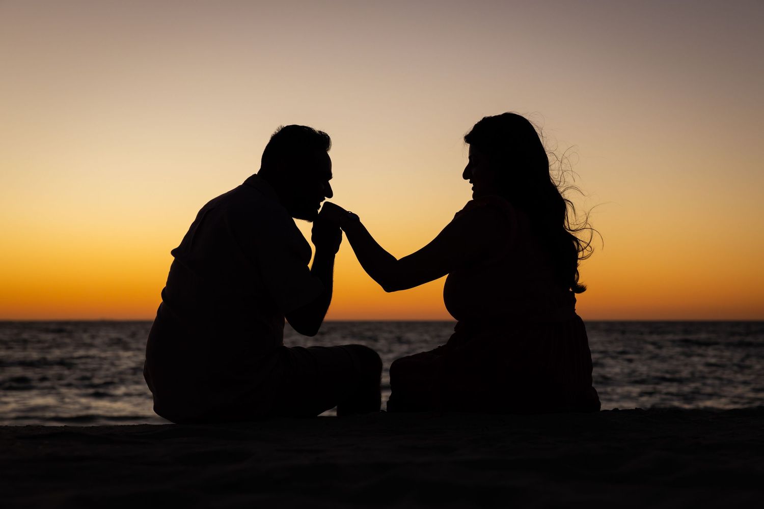 Silhouettes of two people standing together against vibrant orange sunset sky over ocean horizon.