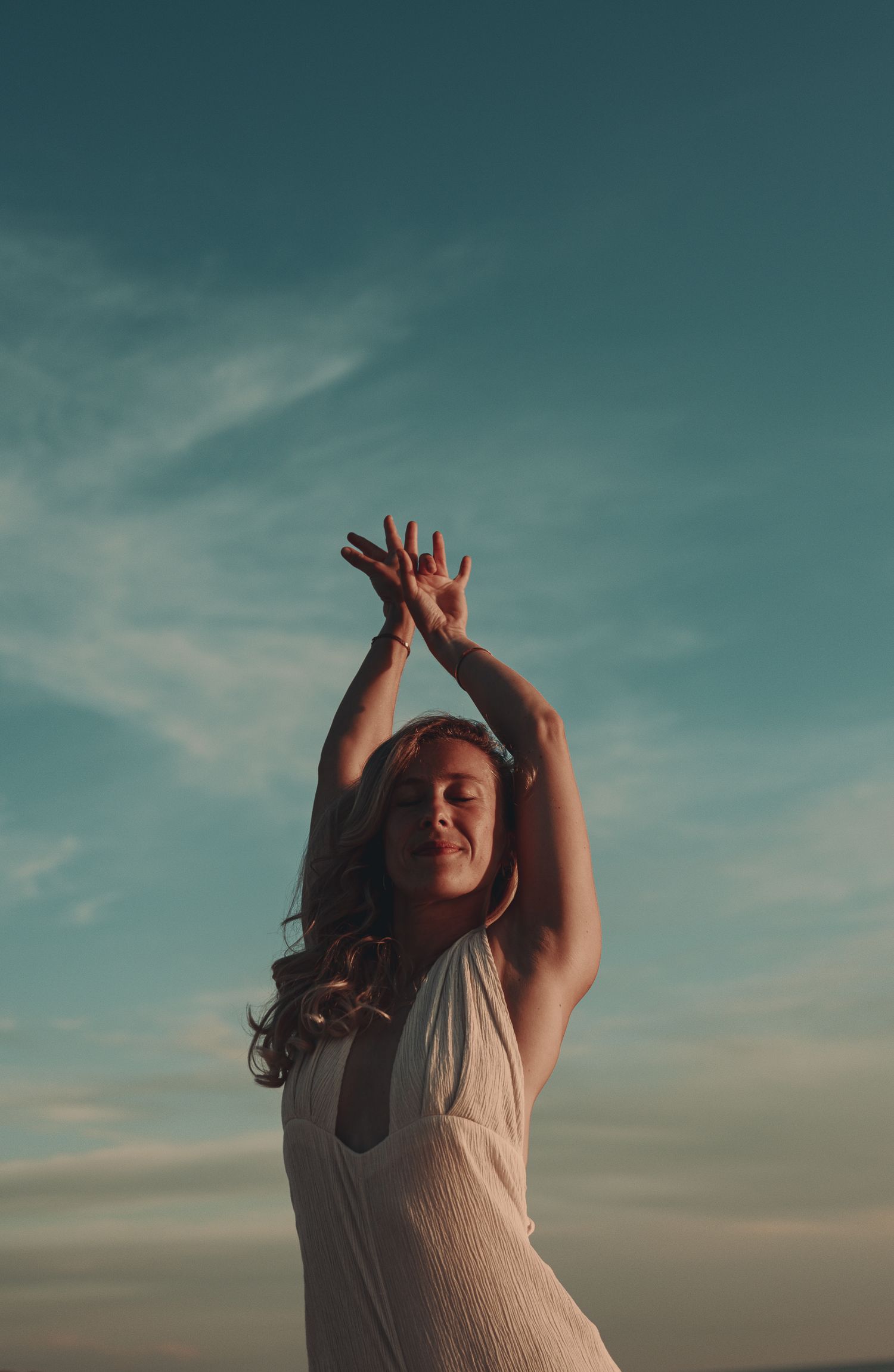 Woman in white dress dancing outdoors with arms raised against a blue sky at sunset.