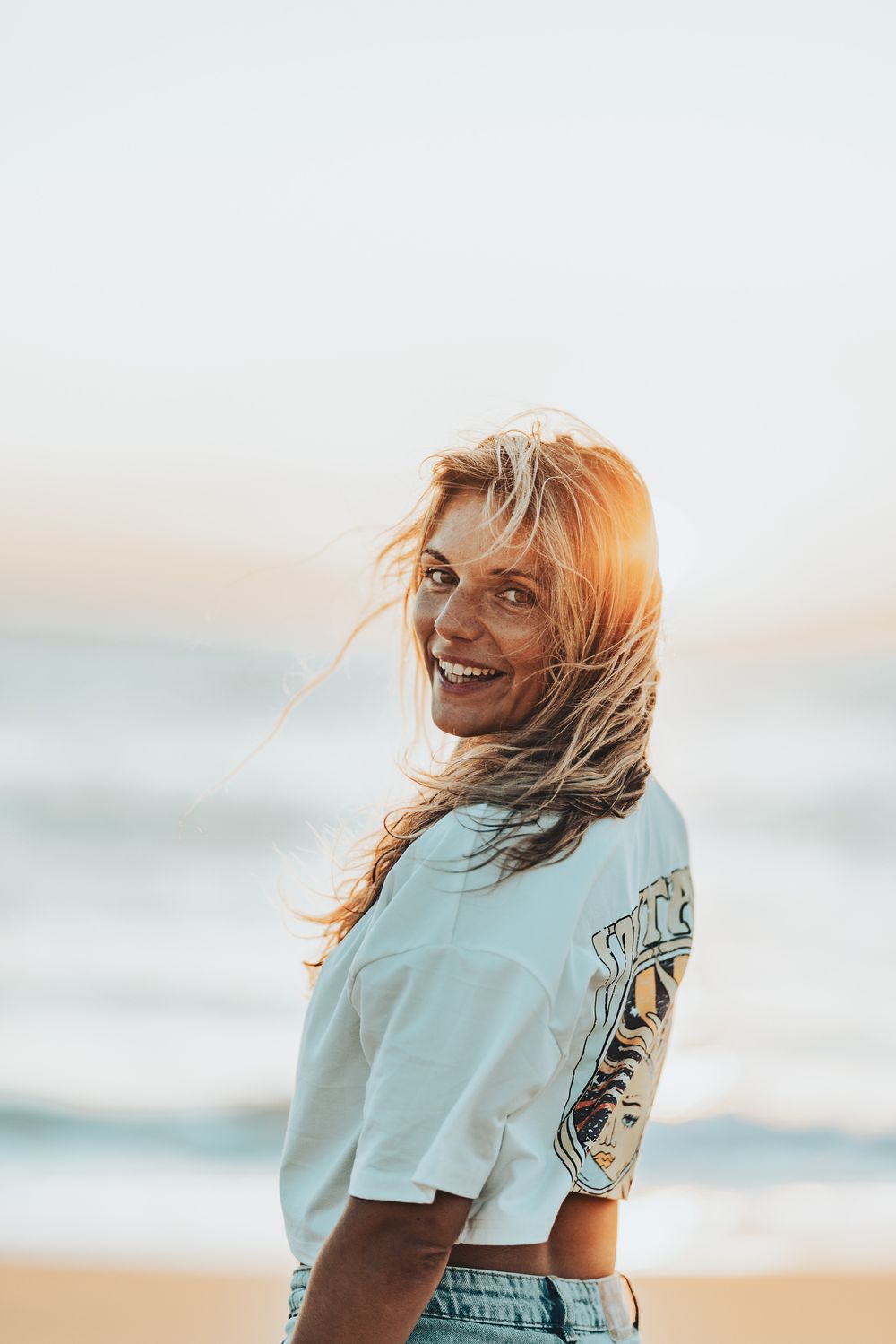 Une personne aux cheveux balayés par le vent sourit au coucher du soleil sur une plage portant un t-shirt bleu clair.