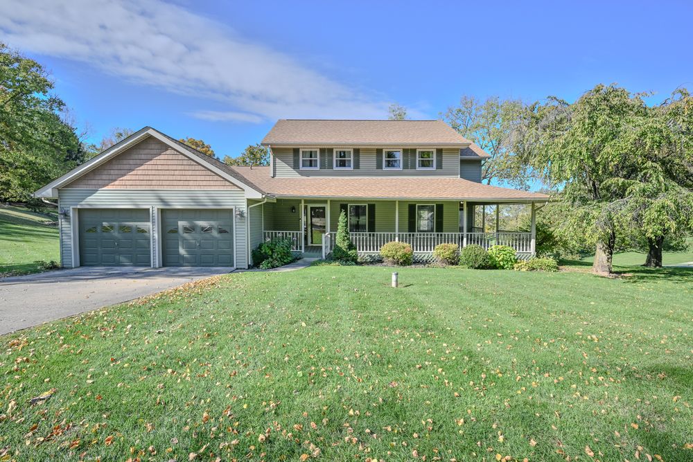 Scenic rural property with green lawn and mature trees surrounds a two-story colonial style home with attached garage.