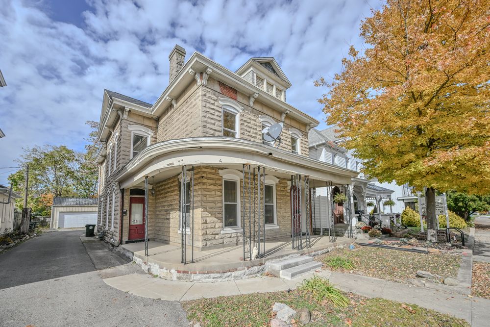 Historic Victorian style stone house with wrap-around porch and dormer windows set against autumn trees and blue sky.