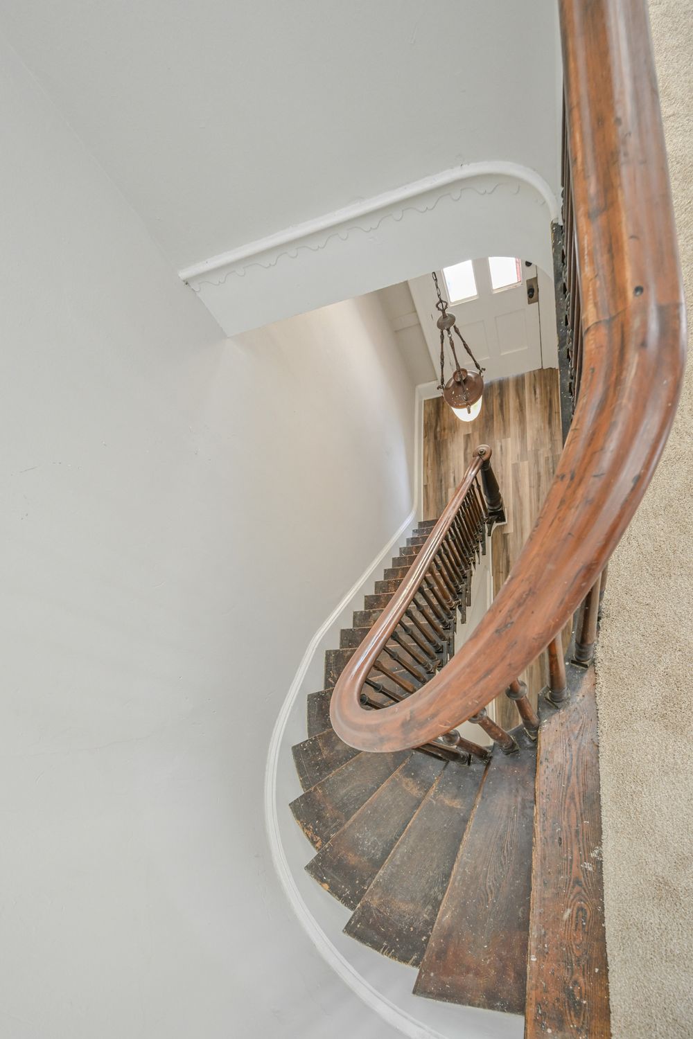 A curved wooden staircase with a white wall and brown handrail photographed from above in a spiral pattern.