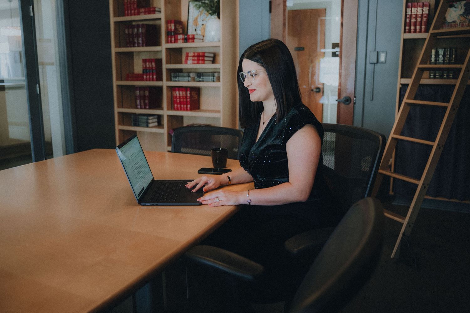 Student working quietly at a wooden desk in library study area with bookshelves visible in background.