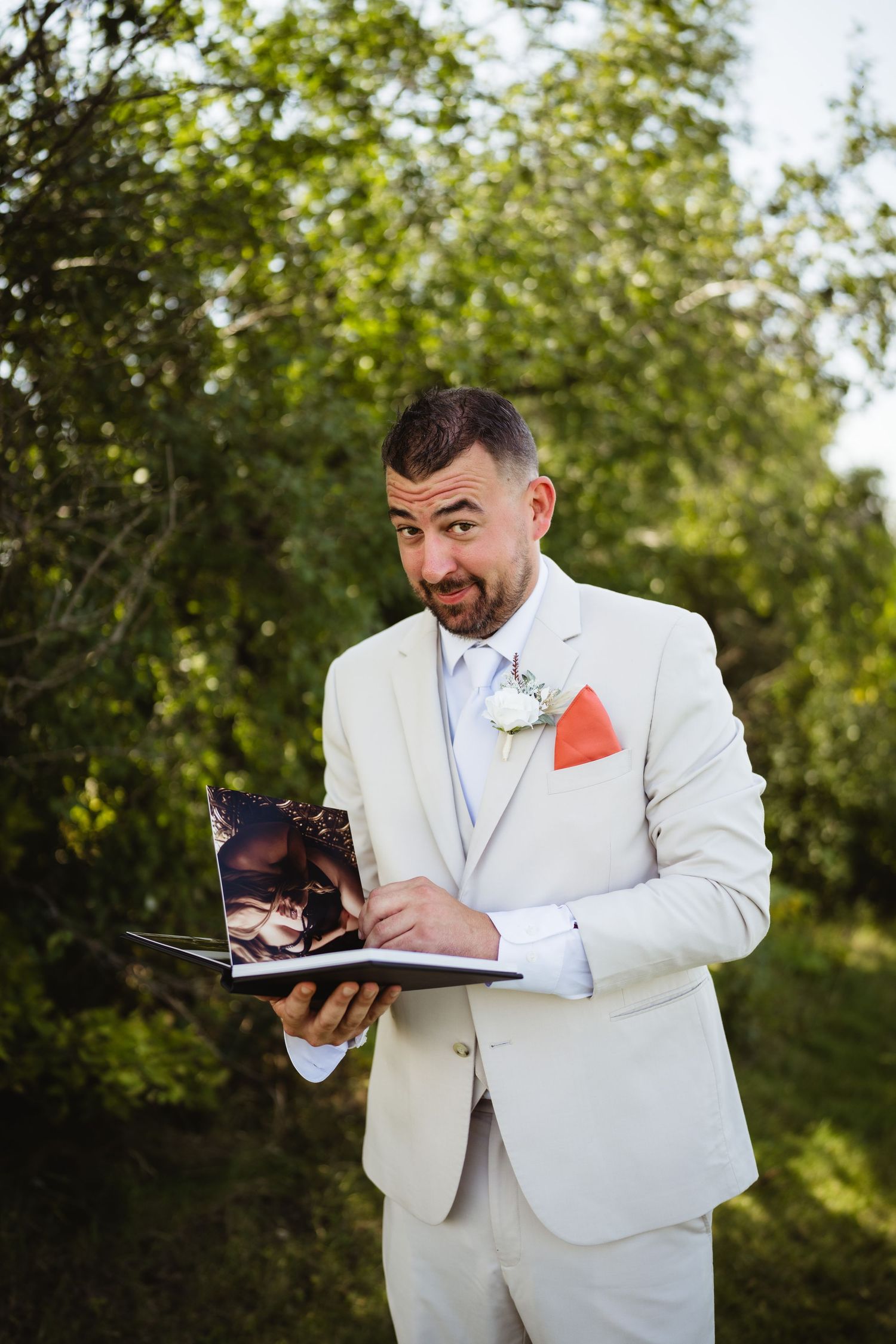 A person in a white suit jacket reads from a small book while standing outdoors in a natural setting.