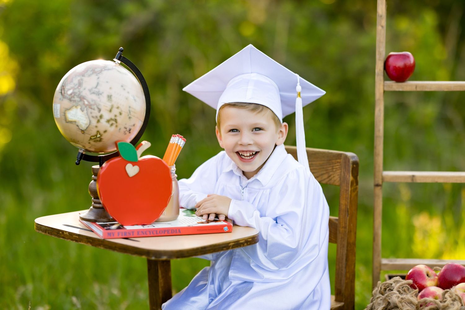 Young student in graduation cap sits at outdoor desk with globe and apple decorations in garden setting.