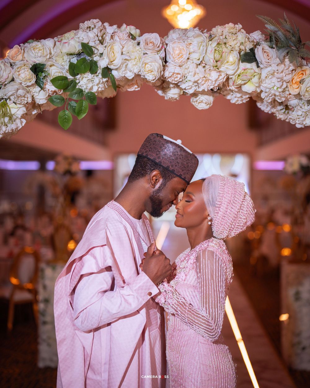 A romantic moment under a floral arch with a couple in coordinated pink outfits sharing an intimate embrace.