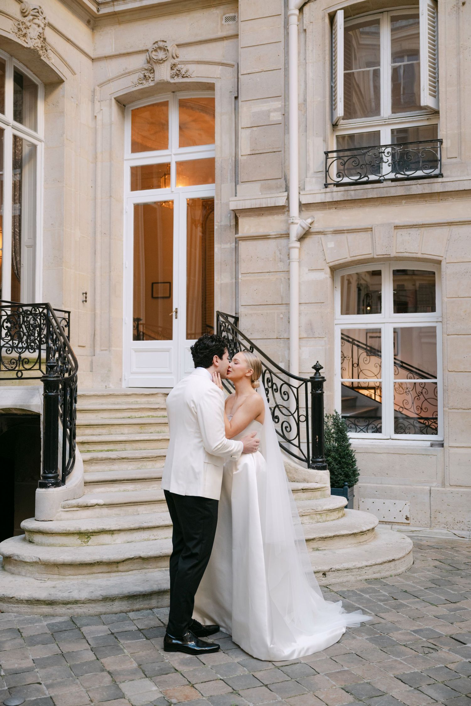 Romantic wedding couple embraces in front of elegant stone building with ornate architectural details and windows.