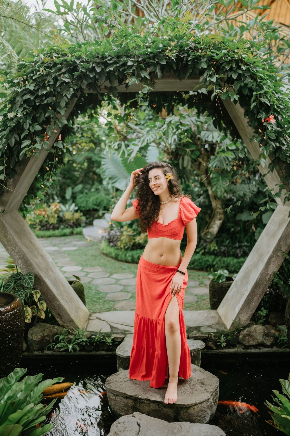 Person in red dress sitting on wooden beams in a tropical garden setting.