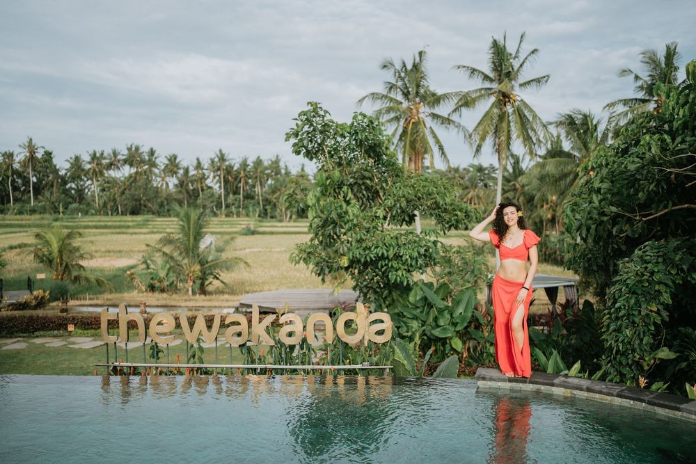 Person in bright red dress walking along waterfront with palm trees in background.