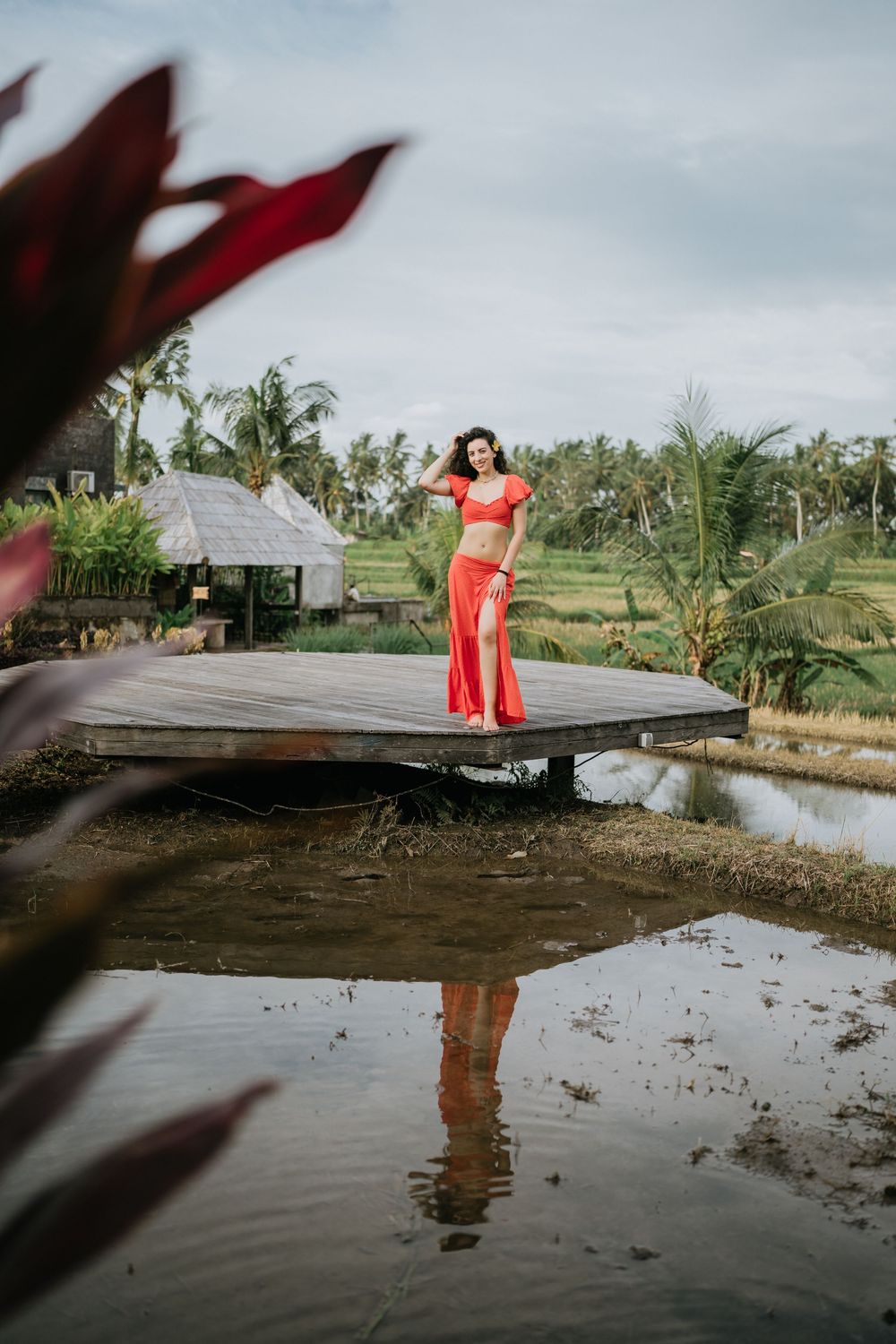 Person in red outfit reflected in still water by wooden dock surrounded by tropical landscape.
