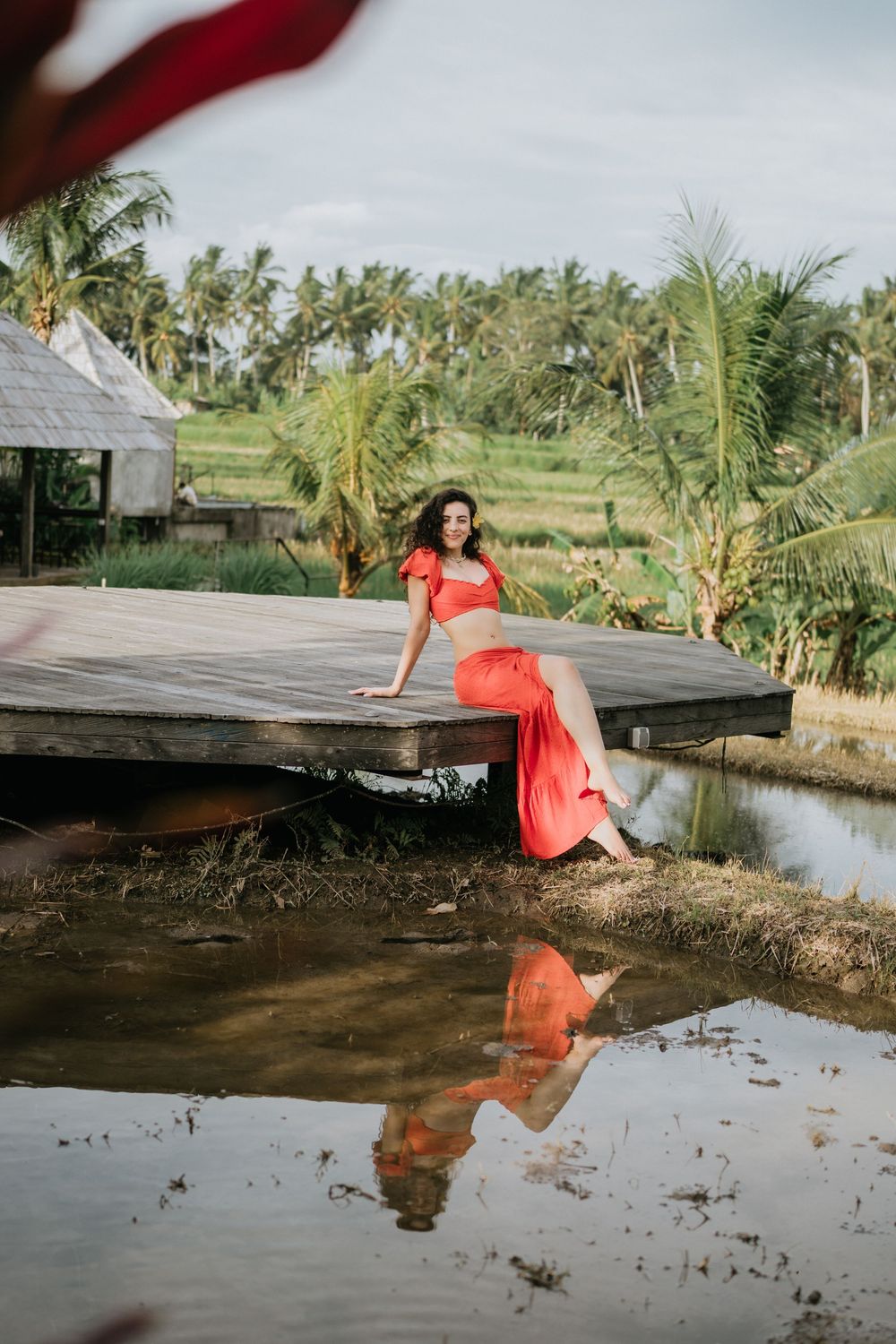 Figure in red dress sitting on wooden dock over reflective water with palm trees in distance.